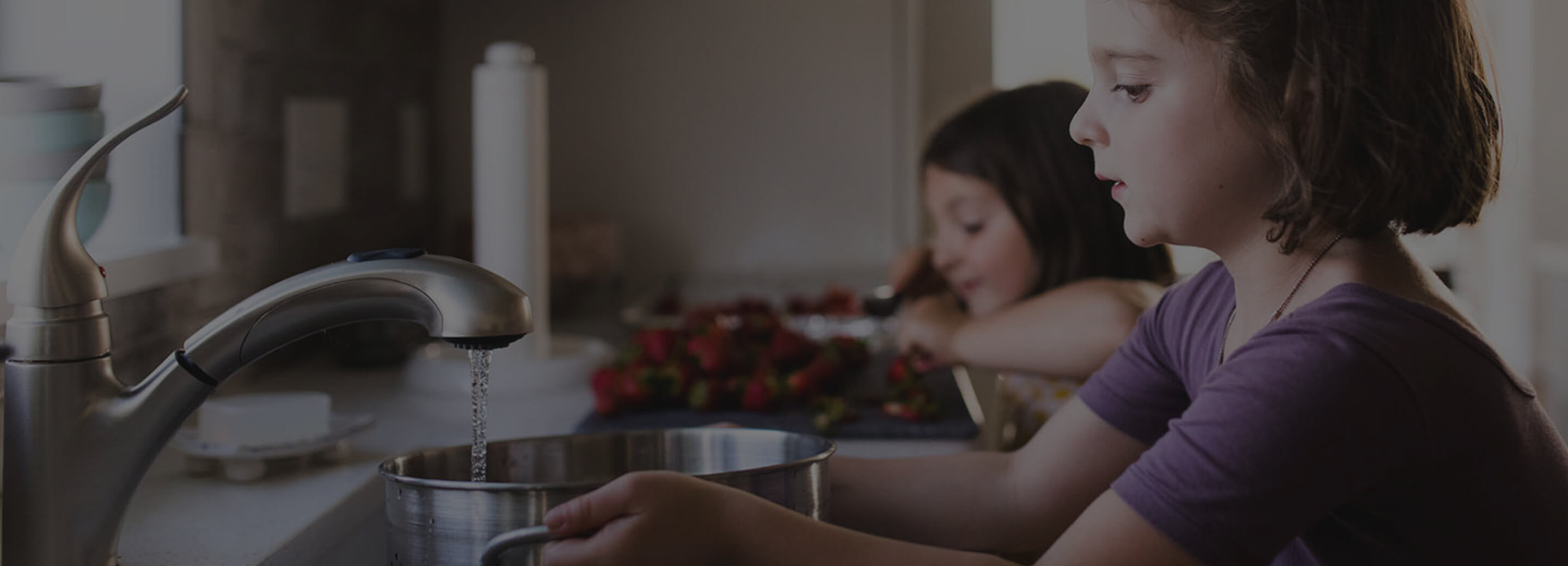 A child filling a pot with water while a child behind her prepares strawberries