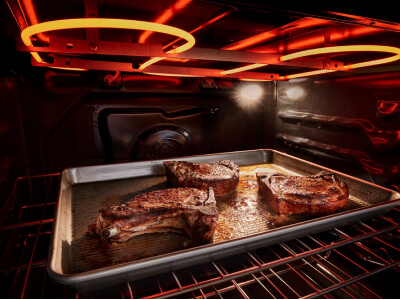 The inside of an oven with a pan of meat in it and the broiler coils lit up