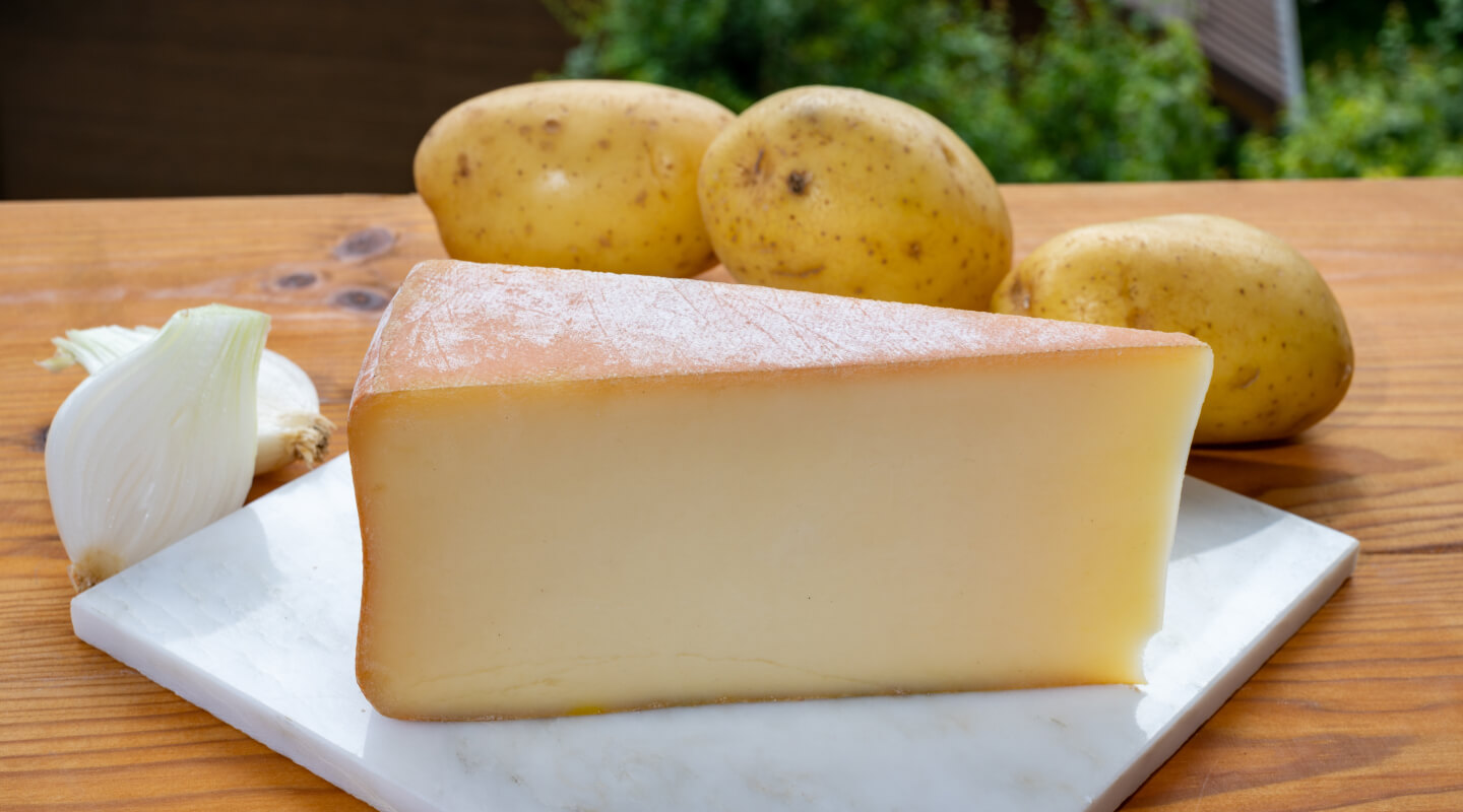 A wedge of cheese on a cutting board with three potatoes behind it