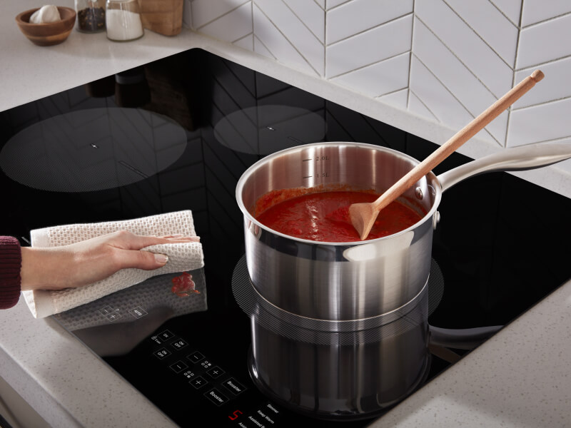 Person wiping cooktop next to burner with saucepan 