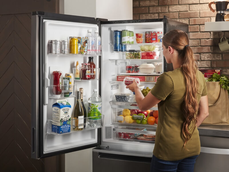 A woman standing in front of an open, bottom-freezer refrigerator  A woman standing in front of an open, bottom-freezer refrigerator