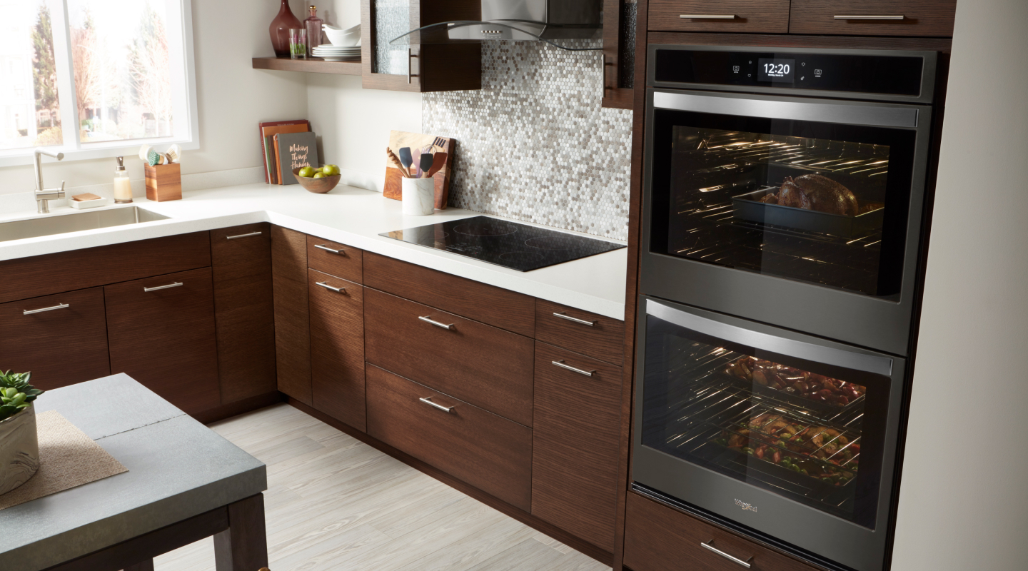 Double wall oven and an electric flat top range in a kitchen with dark wood cabinets and neutral tile backsplash