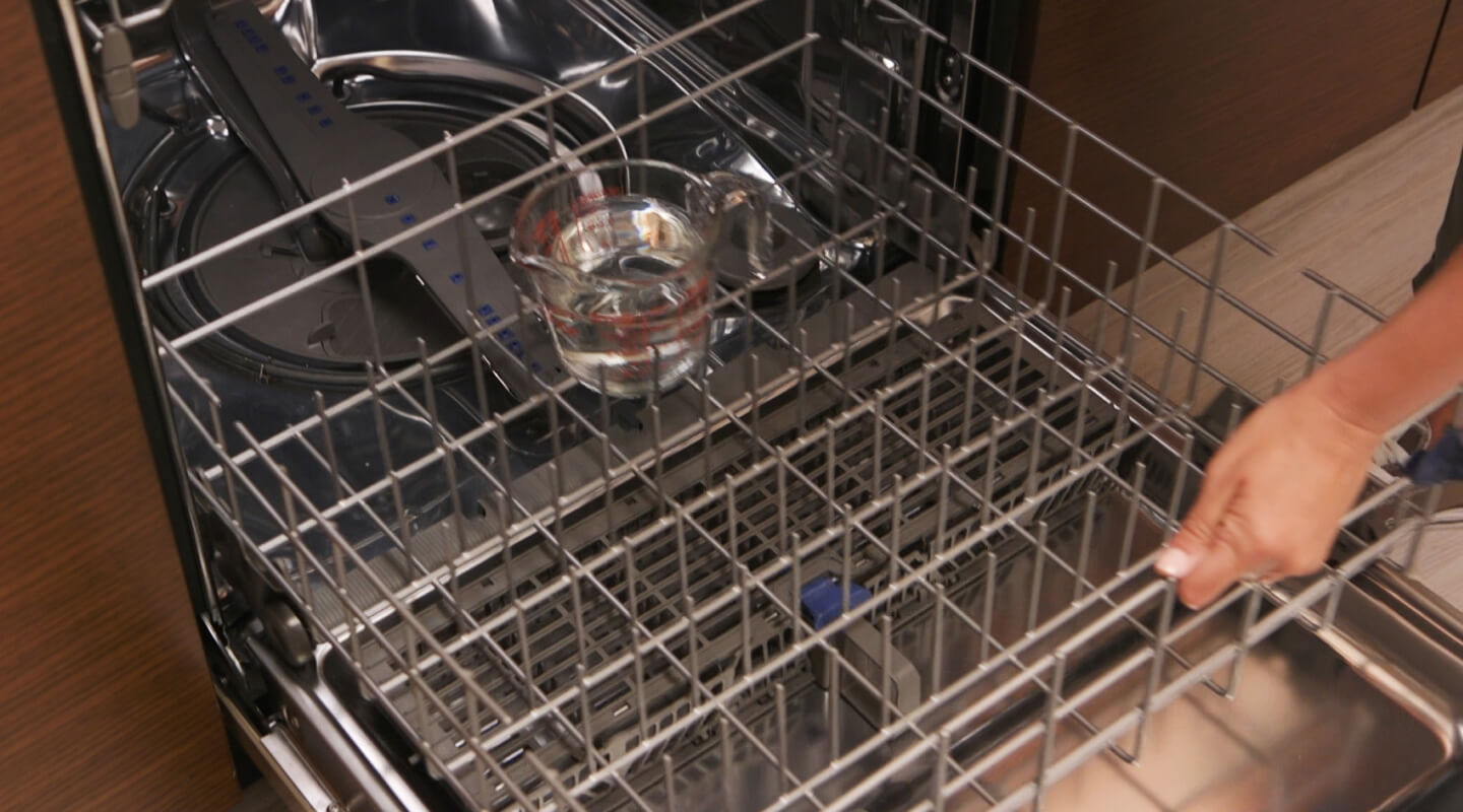A person adding a measuring cup of vinegar to a dishwasher rack