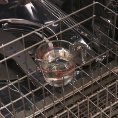 A person adding a measuring cup of vinegar to a dishwasher rack