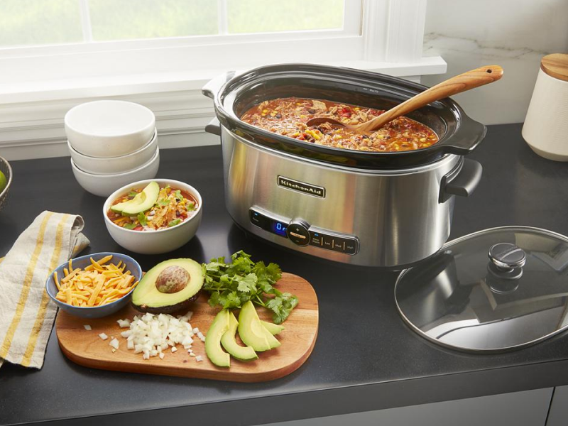 Slow cooker filled with chili next to a cutting board with toppings Slow cooker filled with chili next to a cutting board with toppings