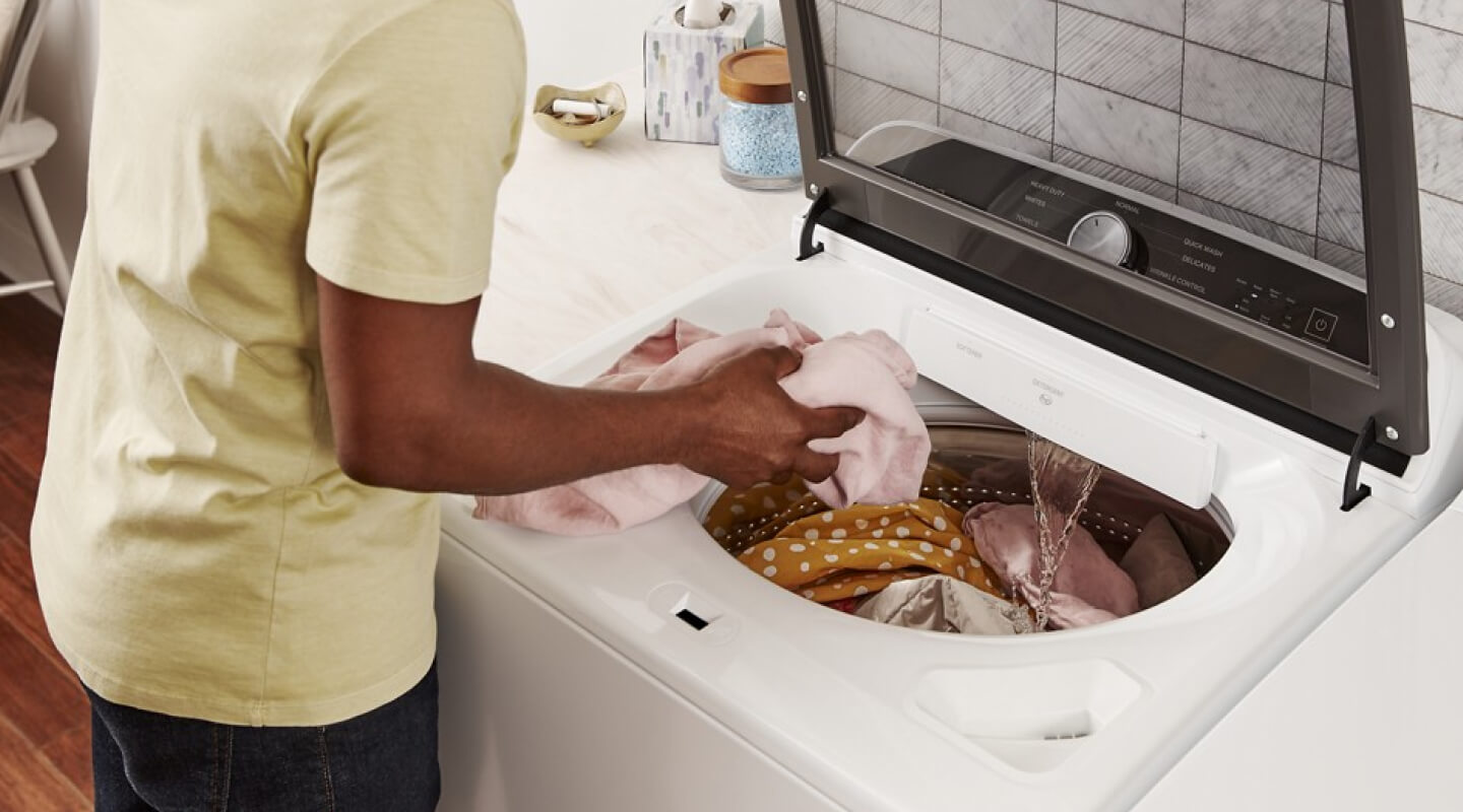 A person putting a pink shirt into a top load washer filling with water