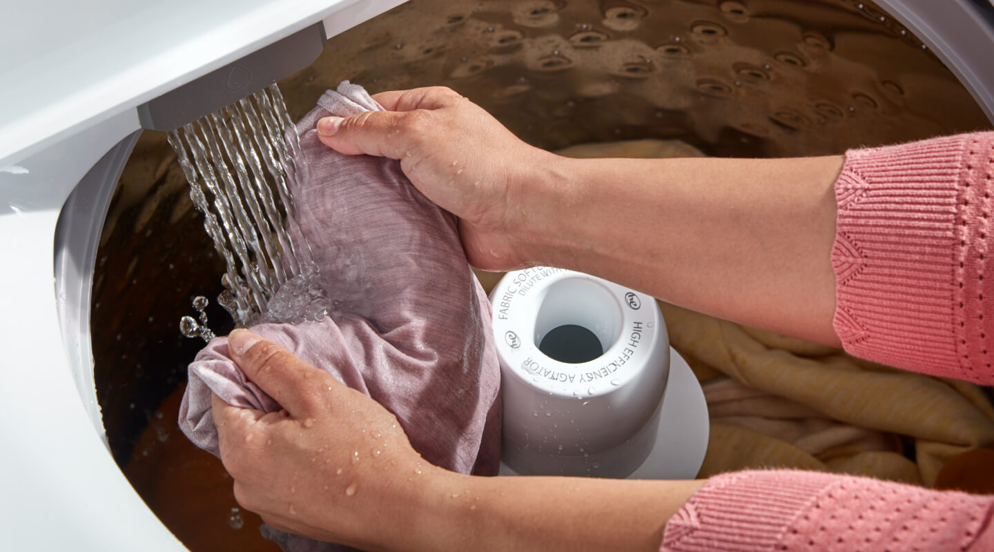 Hands holding a pink garment under a spray of water in a top load washer