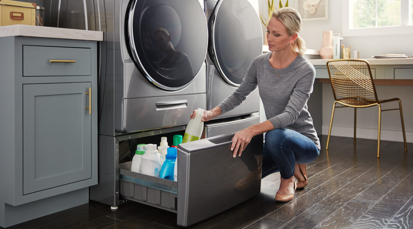 A person putting away detergent in a laundry pedestal that’s under a gray front load washer and next to a front load dryer A person putting away detergent in a laundry pedestal that’s under a gray front load washer and next to a front load dryer