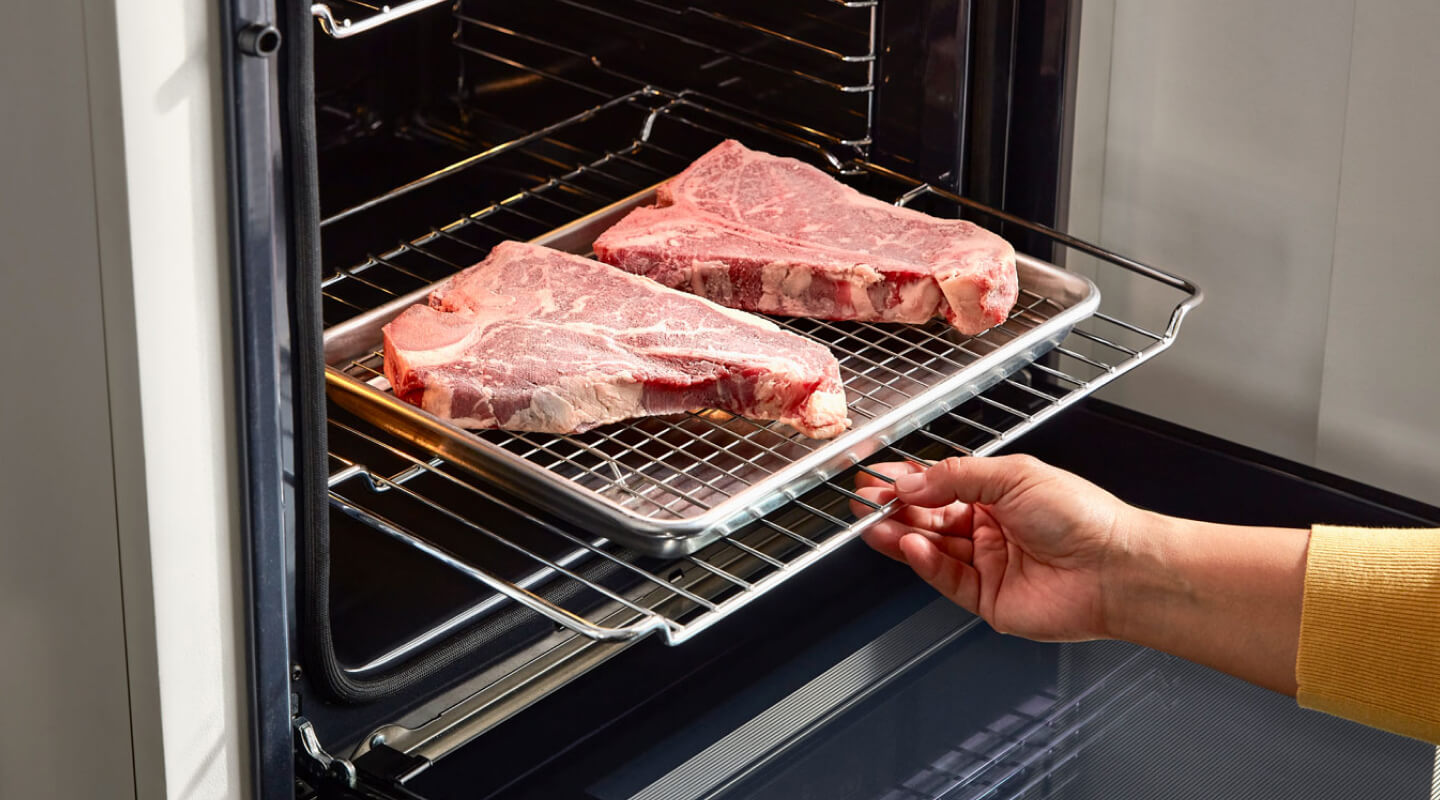 A person sliding a rack of raw steaks into an oven A person sliding a rack of raw steaks into an oven