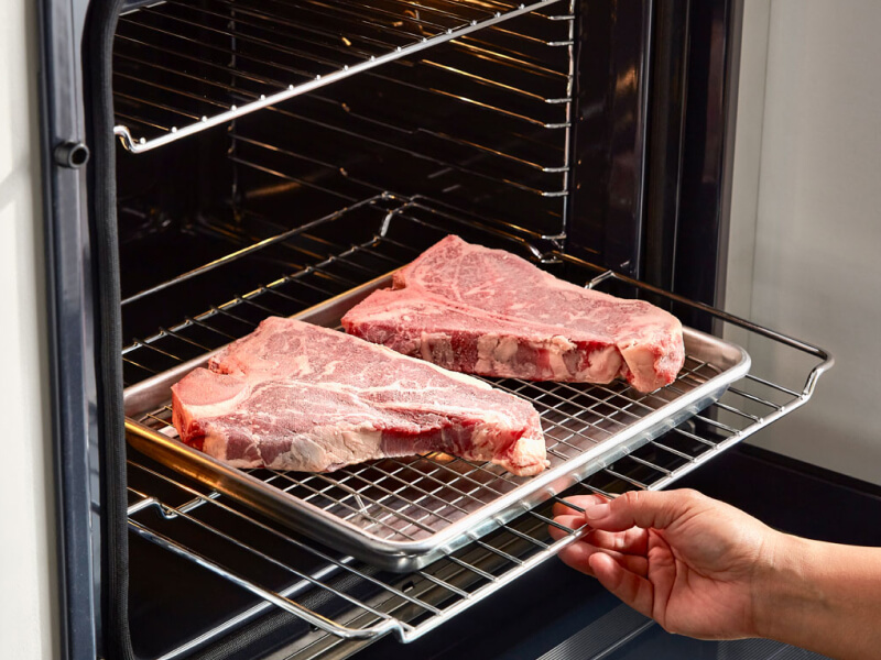 A person sliding a rack of raw steaks into an oven A person sliding a rack of raw steaks into an oven