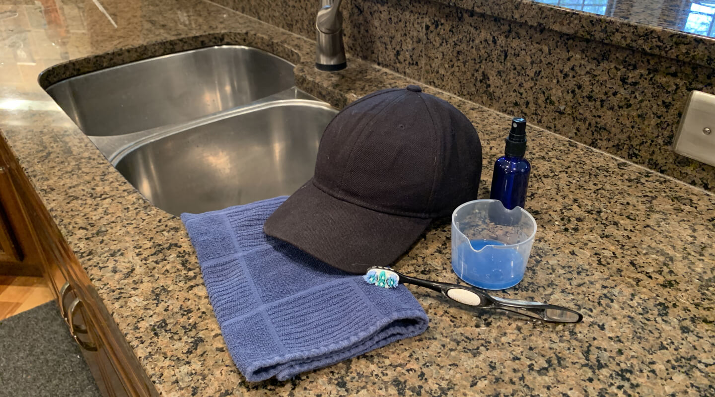 A black cloth baseball cap on a countertop with a toothbrush, towel and cleaning supplies