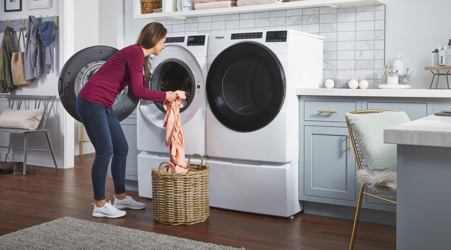 A woman putting laundry into a white Whirlpool® front load washer that sits next to a dryer in a modern laundry room.