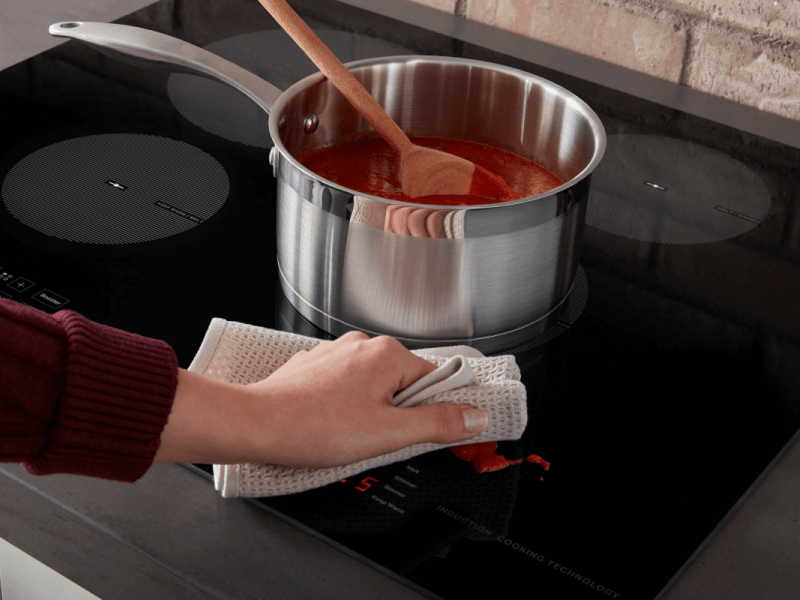 Person wiping sauce splatter off of an induction stovetop Person wiping sauce splatter off of an induction stovetop