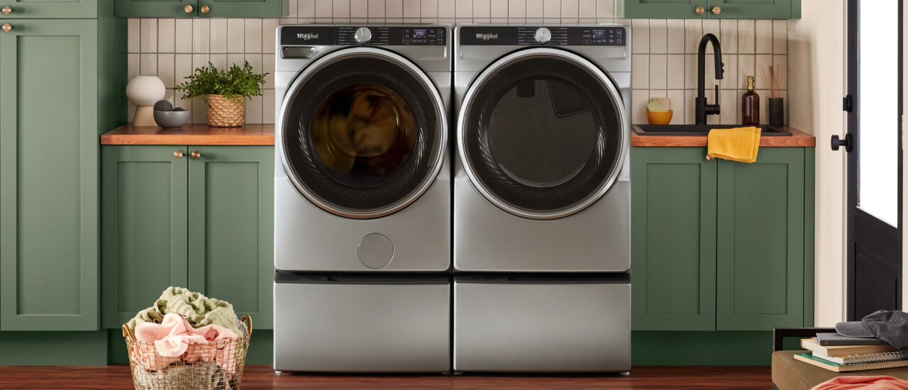 Side-by-side front load washer and dryer on pedestals in a laundry room with green cabinets