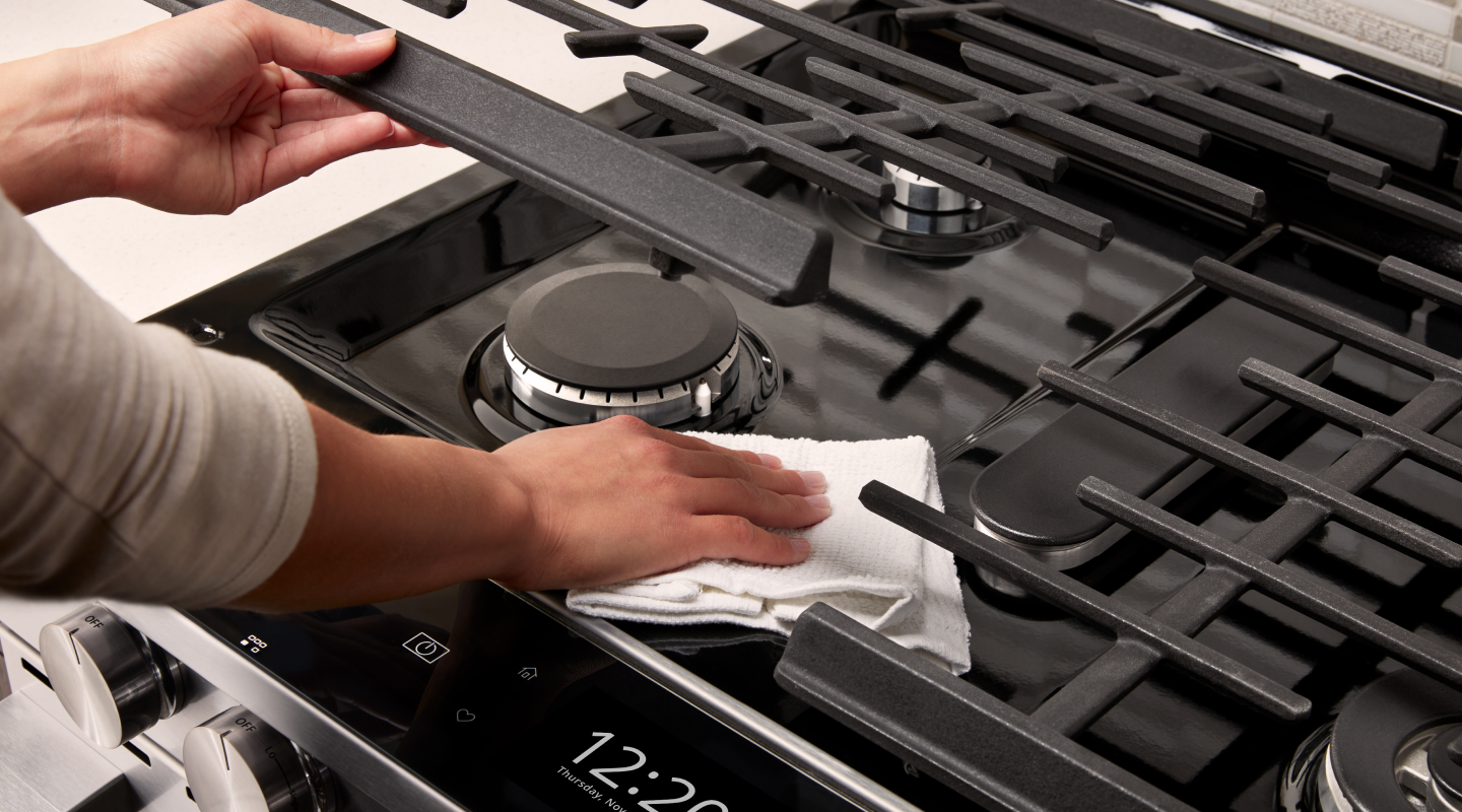 Person wiping underneath a gas cooktop grate Person wiping underneath a gas cooktop grate