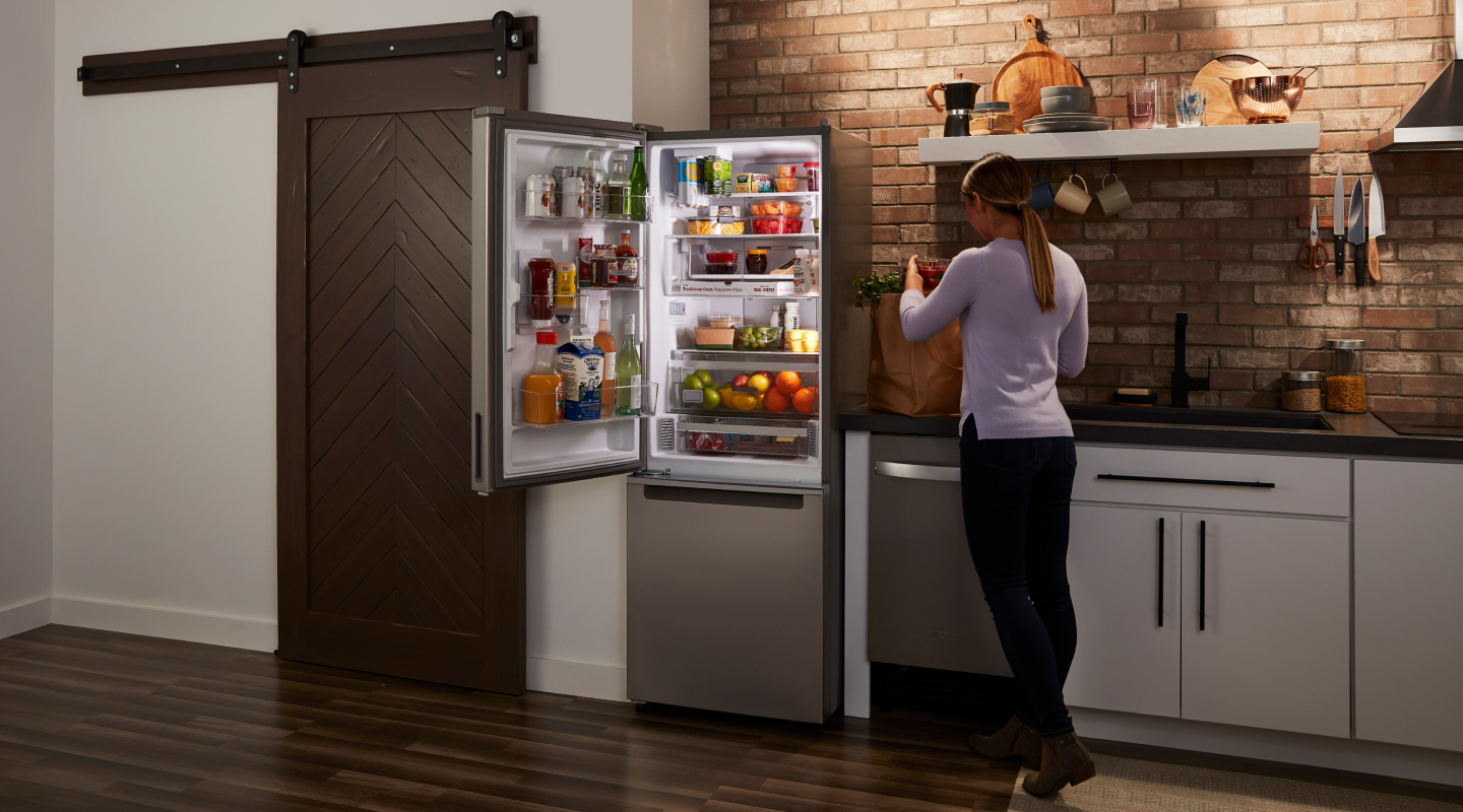A woman in a kitchen by an open refrigerator stocked with food