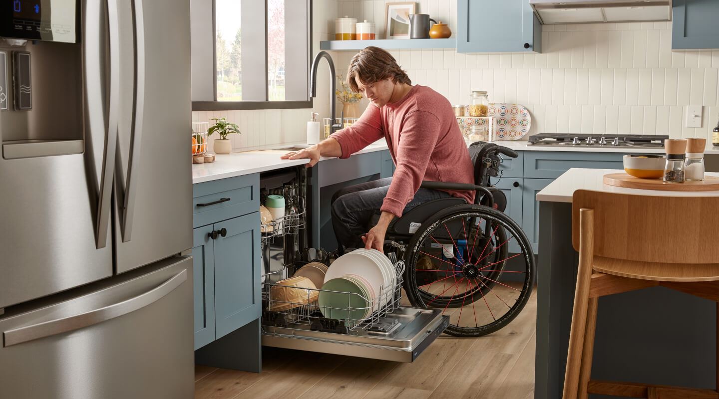 A person in a wheelchair reaching for dishes in an open built-in dishwasher that’s placed in a kitchen with powder blue cabinets and white countertops