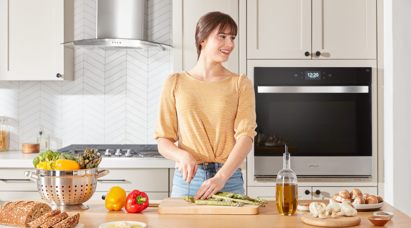 A person chopping asparagus on a wooden cutting board placed on a kitchen island with a cooktop in the background behind her A person chopping asparagus on a wooden cutting board placed on a kitchen island with a cooktop in the background behind her