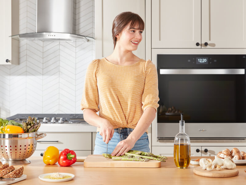 A person chopping asparagus on a wooden cutting board placed on a kitchen island with a cooktop in the background behind her A person chopping asparagus on a wooden cutting board placed on a kitchen island with a cooktop in the background behind her