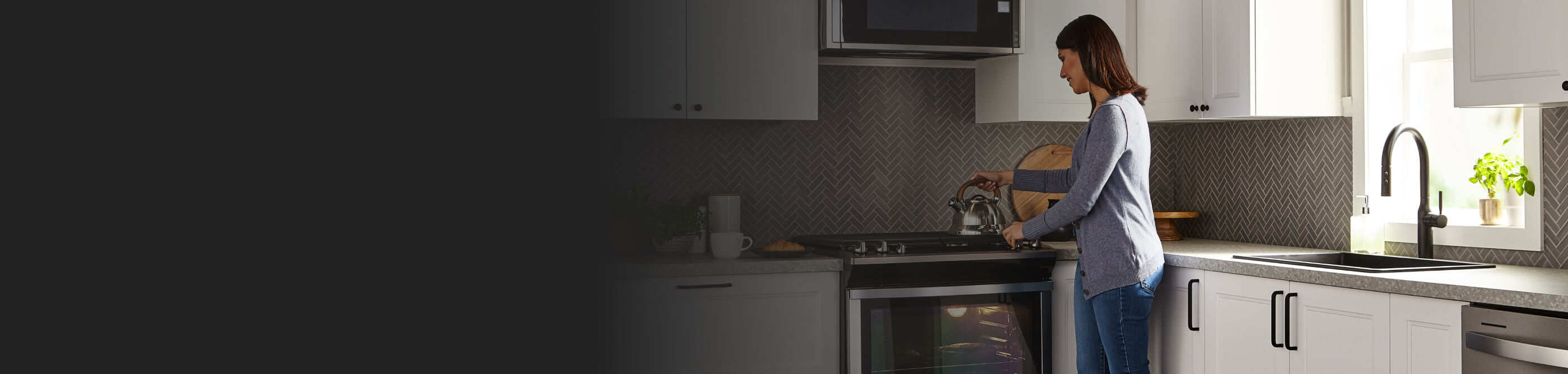 Person standing over a gas range with a hand on a tea kettle that’s resting on a stovetop in a kitchen with white cabinetry