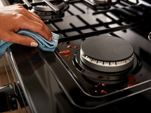Closeup of a hand holding a blue cloth, wiping marina sauce splatters from a black gas stovetop surface