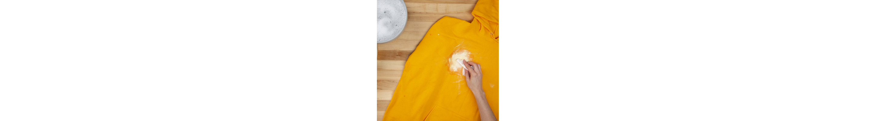 Person using a spoon to scrape off dried white paint from a yellow sweatshirt